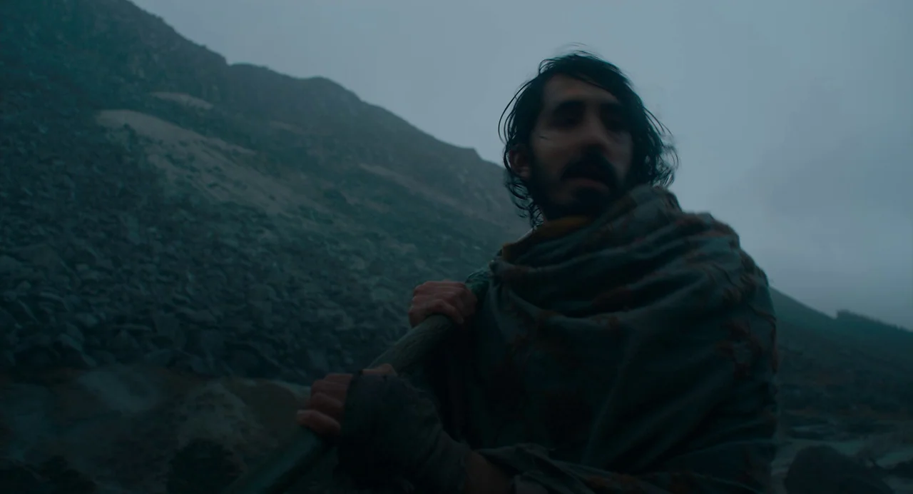 The Green Knight (2021) movie still: medium — A rocky, mountainous landscape under a dense, overcast, grey sky. A man in a heavy textured cloak…