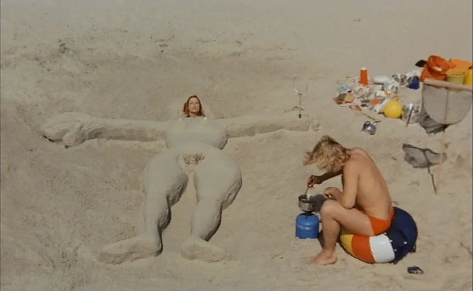 Turkish Delight (1973) movie still: wide — A sunny sandy beach features a large, human-shaped sand sculpture containing a woman's head. Beside the…