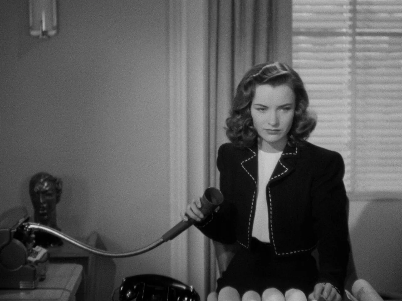 Phantom Lady (1944) movie still: medium — A 1940s workspace features a desk with a dictaphone and venetian blinds behind. A woman in a dark stitched…