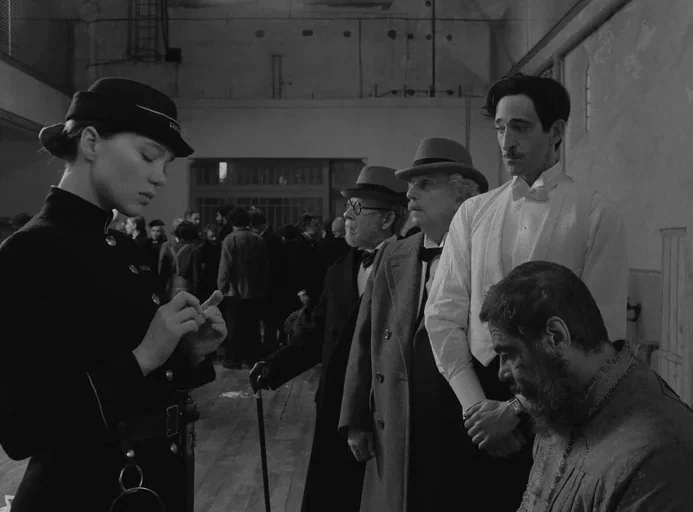 The French Dispatch (2021) movie still: medium — A group of people stands in a sparsely furnished, large interior room with wood floors. A woman in…