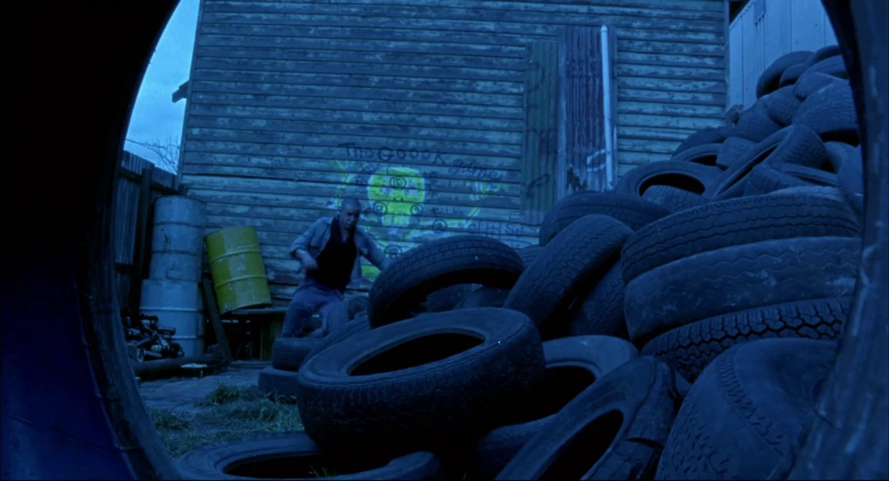 Romper Stomper (1992) movie still: wide — A cluttered outdoor storage area features a large stack of worn rubber tires. A man stands near a…