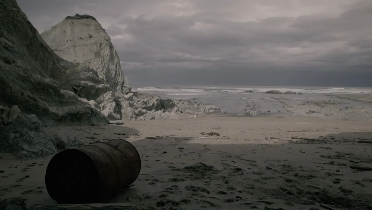 Hondar 2050 (2018) movie still: establishing — A rugged beach features a large rusted metal drum in the foreground. A rocky cliff rises on the left…
