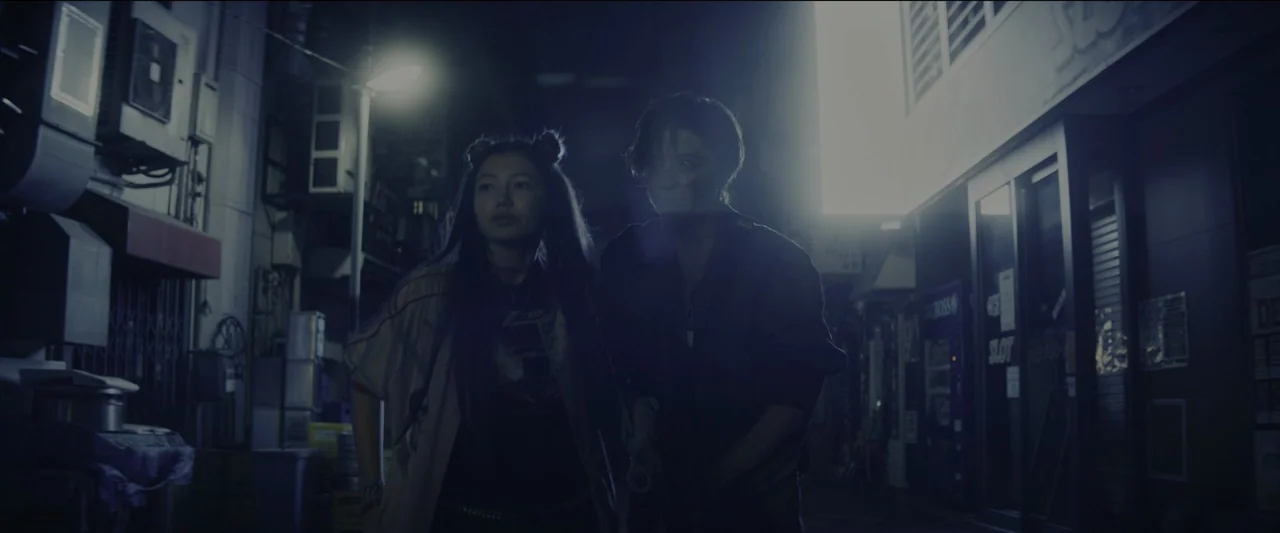 Kate (2021) movie still: medium — A narrow urban street at night with metal security gates and closed storefronts. Two young people walk together…