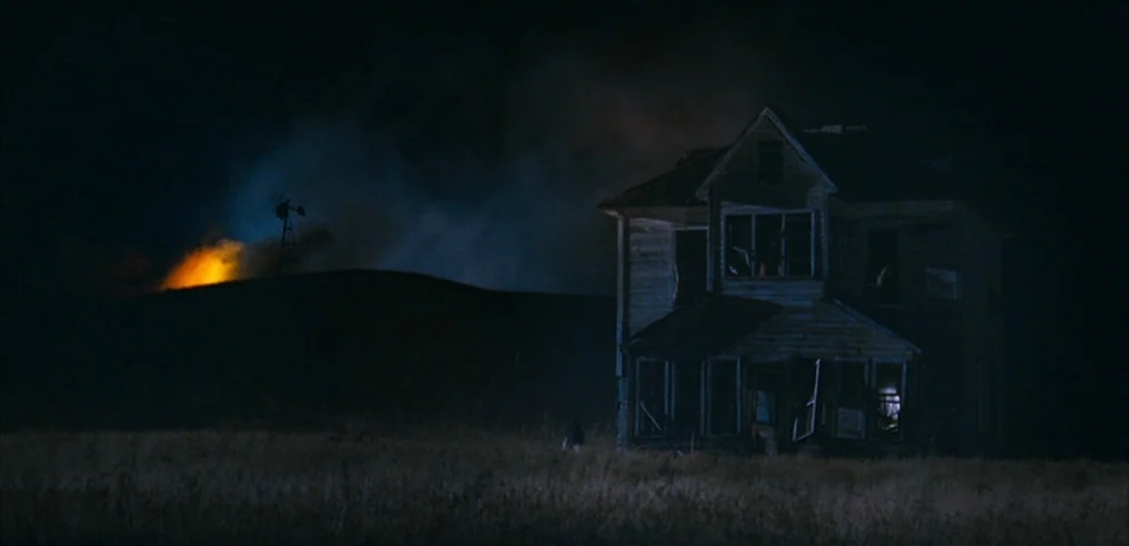 Tideland (2005) movie still: establishing — A dilapidated wooden farmhouse stands dark in a wide field at night. A distant hill is lit by the orange…