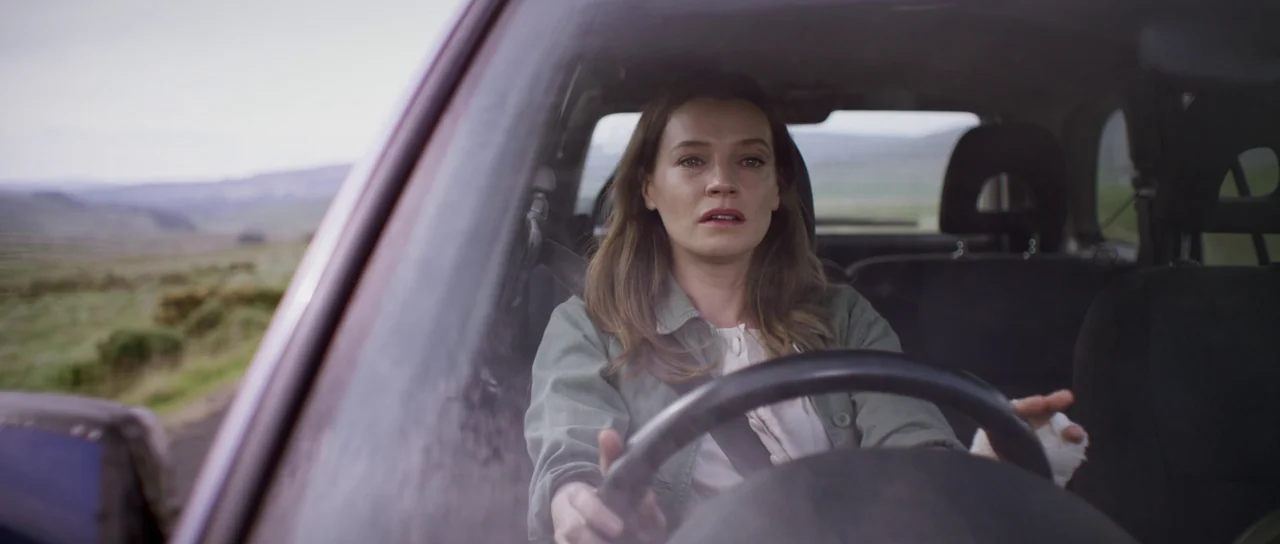 A Dark Song (2016) movie still: medium — A woman drives a car along a road through a rural landscape. She holds the steering wheel with one hand…
