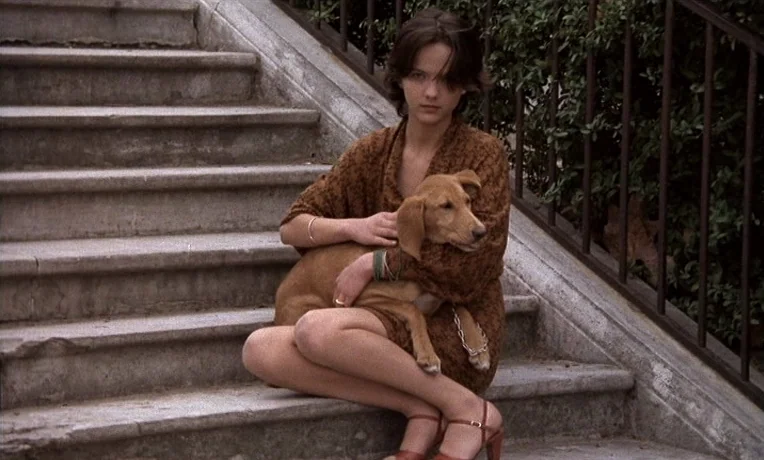 The Man Who Loved Women (1977) movie still: medium — A young woman sits on a set of light-colored stone steps outdoors. She holds a small brown dog…