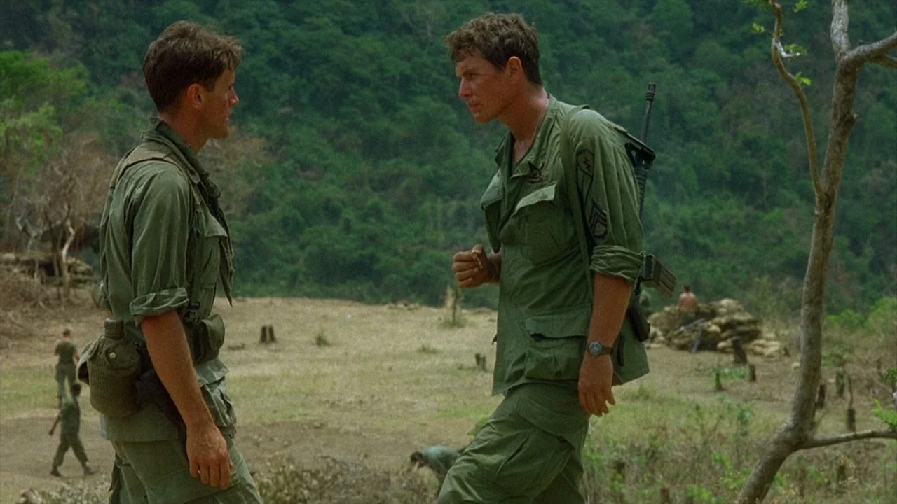 Platoon (1986) movie still: medium — A cleared area of jungle with sparse trees and scattered vegetation. Two soldiers in military gear stand facing…