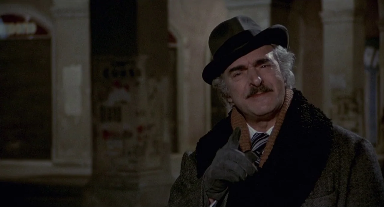 Amarcord (1973) movie still: medium — A man stands outdoors at night near a blurred stone building with poster residue. He wears a formal overcoat…