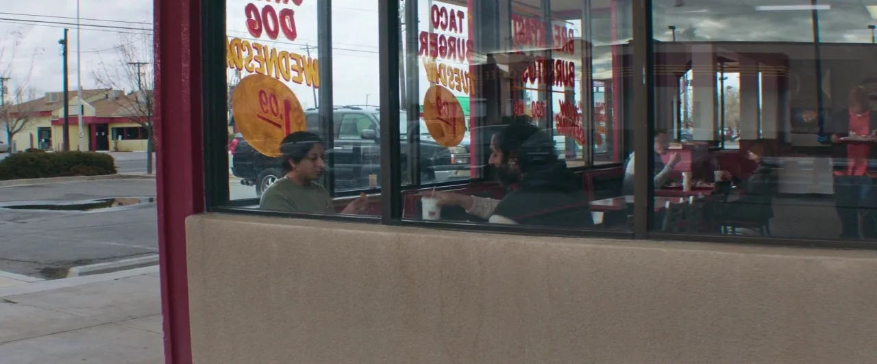 The Long Dumb Road (2018) movie still: medium — A fast food establishment viewed from the sidewalk through large plate glass windows. Two men sit at…