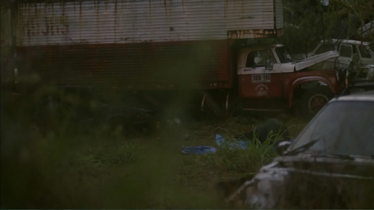 Boy (2010) movie still: wide — A cluttered outdoor scrapyard contains several abandoned vehicles including a large rusted semi-truck. Weeds and tall…