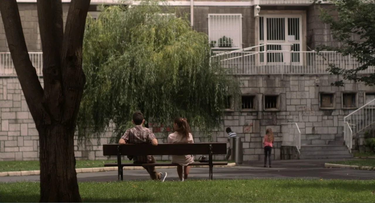 Newton's Third Law (2017) movie still: fullbody — An outdoor park with a stone wall building and a willow tree behind them. A teenage boy and a…