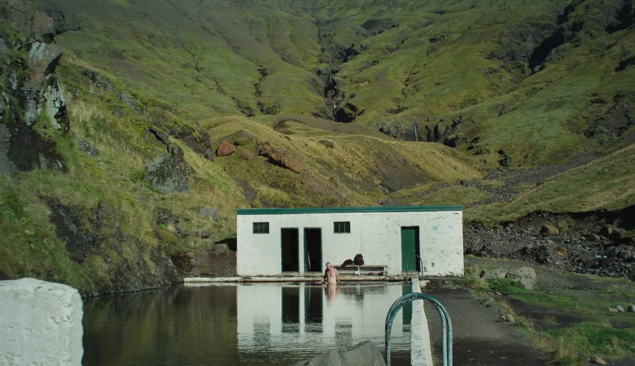 Land Ho! (2014) movie still: wide — A man sits in a concrete geothermal pool nestled at the base of a green mountain. The pool is bordered by a…