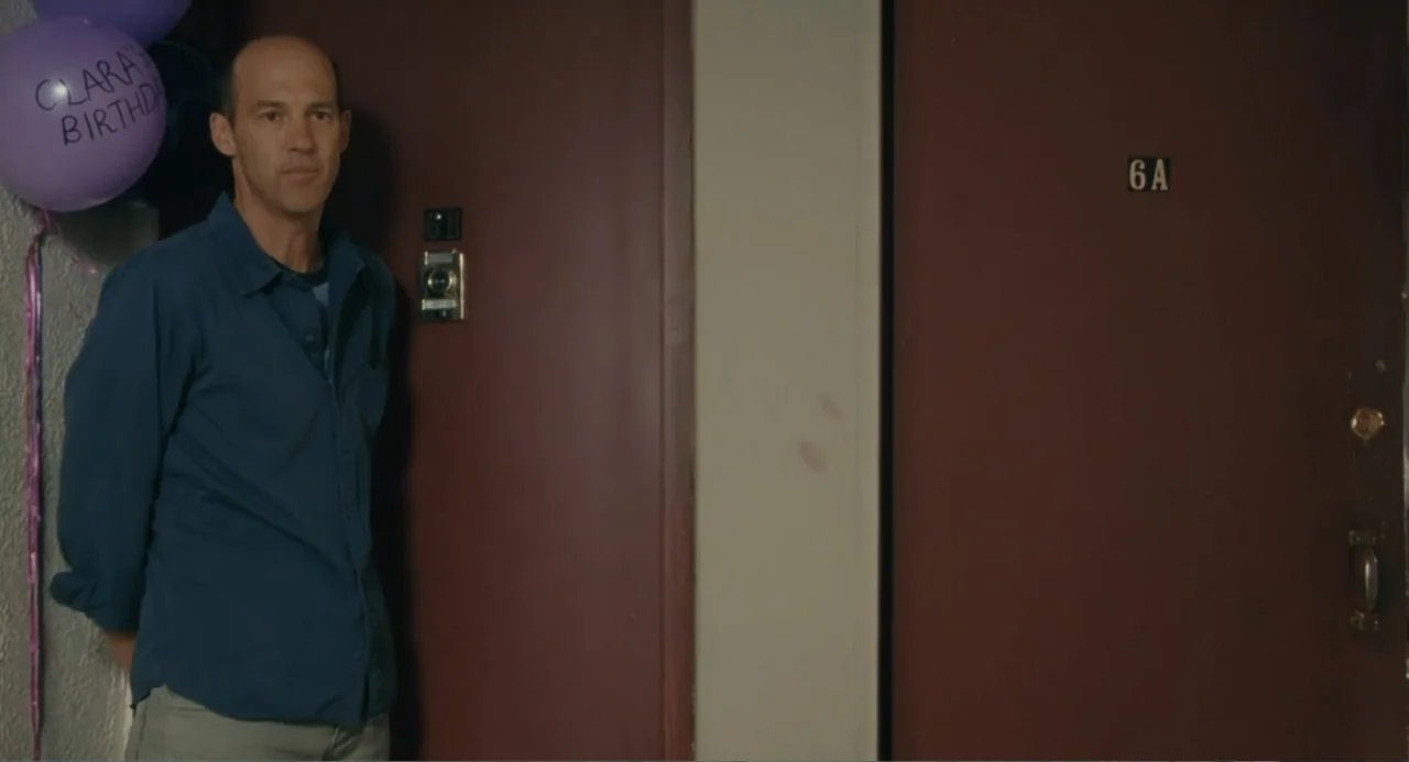 Motherhood (2020) movie still: medium — An apartment hallway features two brown doors labeled 6A and 6B. A bald man stands in front of a door while…