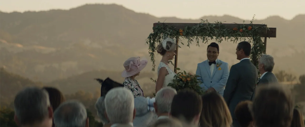 Destination Wedding (2018) movie still: medium — An outdoor wedding ceremony takes place on a hillside overlooking hazy rolling mountains. The bride…