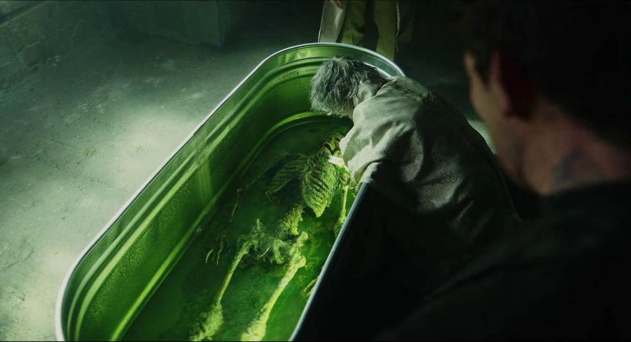 Wake Up Dead Man (2025) movie still: medium — An elderly person leans over a large metal basin filled with thick green liquid. A submerged skeleton…