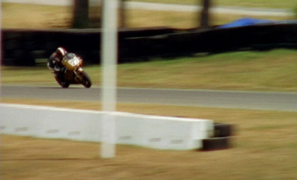 The Brown Bunny (2003) movie still: wide — A motorcycle racer leans into a turn on a paved track with dry grass. White concrete barriers line the…