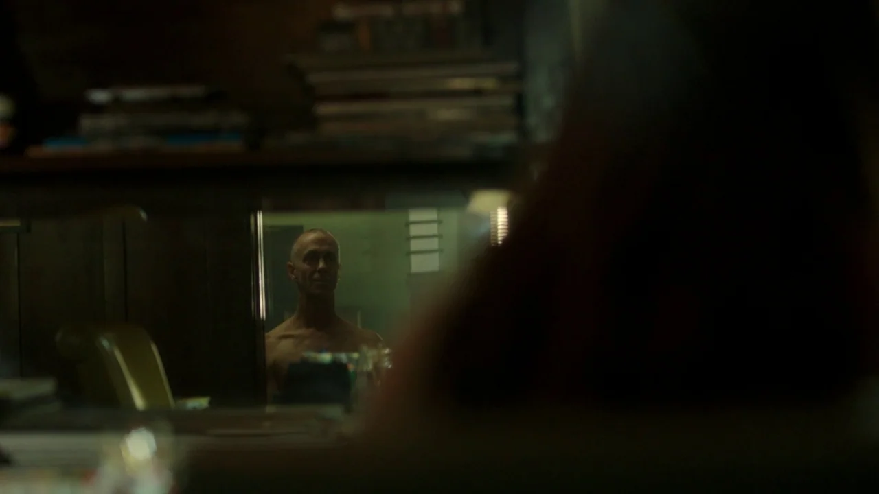 The Pact II movie still: medium — A dark office setting where a man is reflected in a pane of glass. The man appears shirtless and stares toward the…