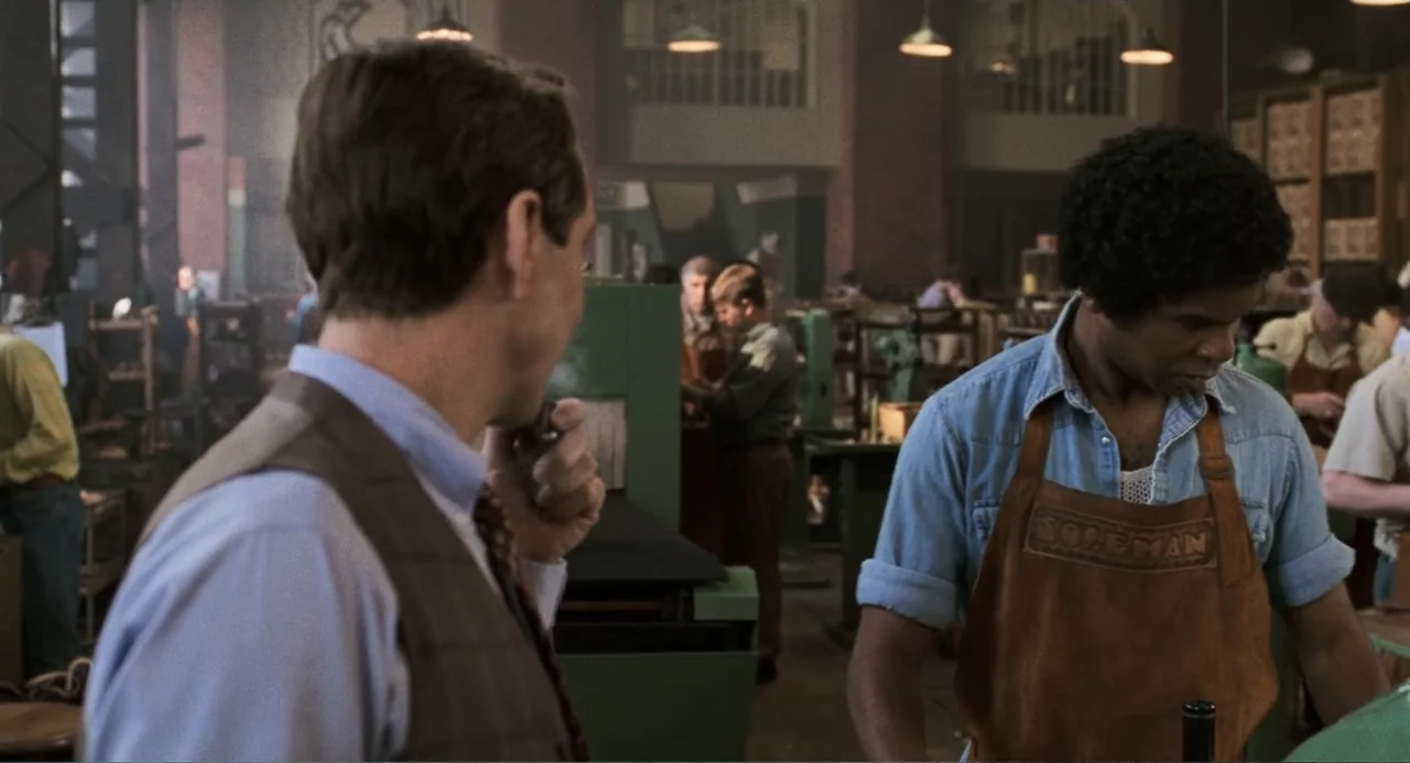 Jumanji (1995) movie still: medium — A cluttered industrial factory floor contains heavy green machinery and multiple workers. A man in a vest…
