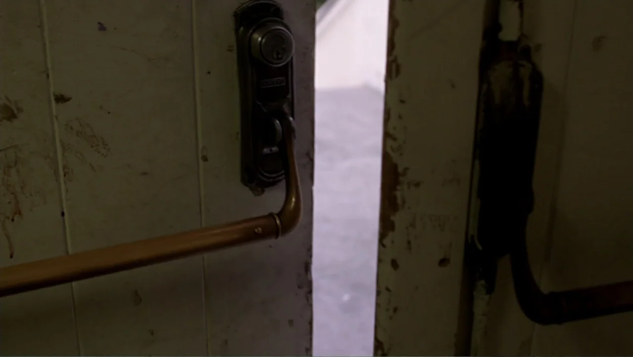 The Gallows (2015) movie still: closeup — A weathered white interior door features a metallic crash bar and a locking mechanism. The door is…