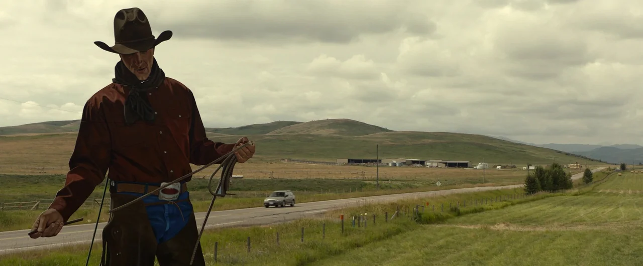 Ghostbusters: Afterlife (2021) movie still: medium — A man dressed as a cowboy stands in a grassy field beside a road. A car travels along the…