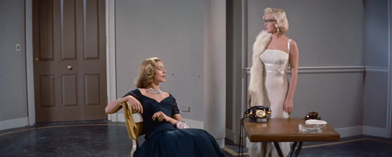 How to Marry a Millionaire movie still: fullbody — An interior space with grey walls, a closed door, and a small wooden table. One woman sits in a…