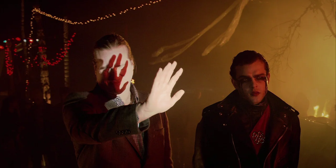 Twixt (2011) movie still: medium — A dark outdoor space is illuminated by warm firelight and distant string lights. One man holds up his palm while…