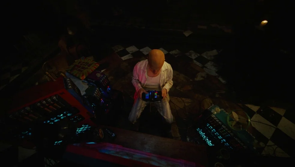 The Zero Theorem (2013) movie still: wide — A dark space features a black and white checkered floor and various glowing equipment. A bald man in…
