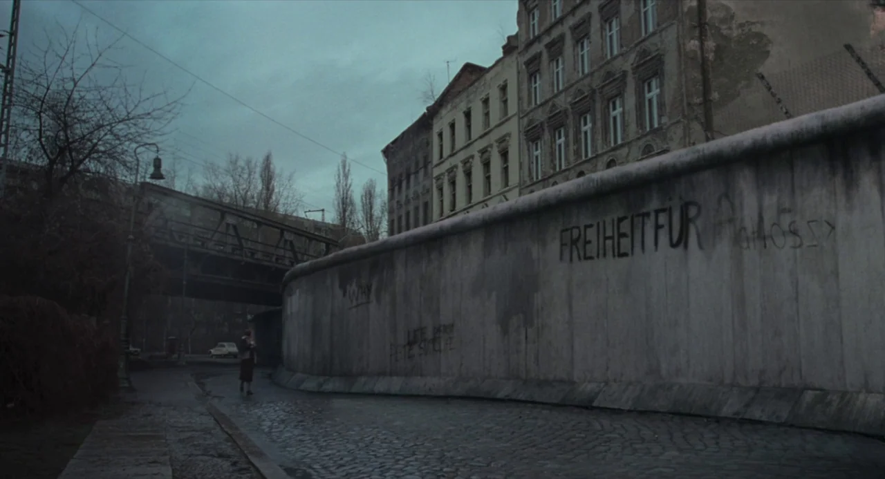 Suspiria (2018) movie still: wide — A tall concrete wall with graffiti stands beside a wet cobblestone street in Berlin. The overcast sky hangs over…