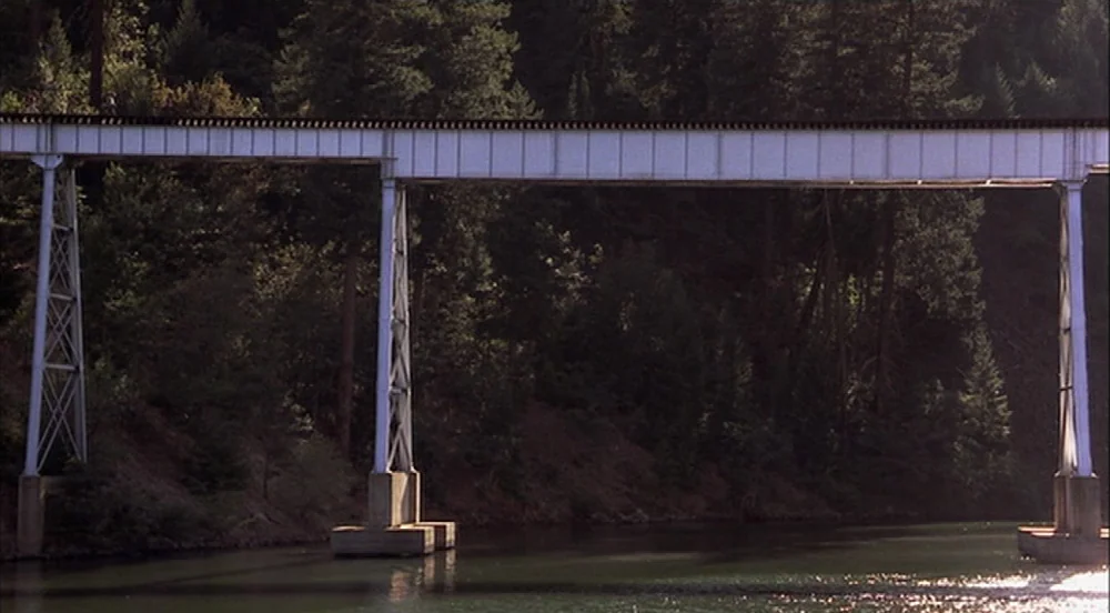 Stand By Me (1986) movie still: wide — A metal railway trestle bridge spans across a wide river flanked by dense forest. The bridge is elevated on…