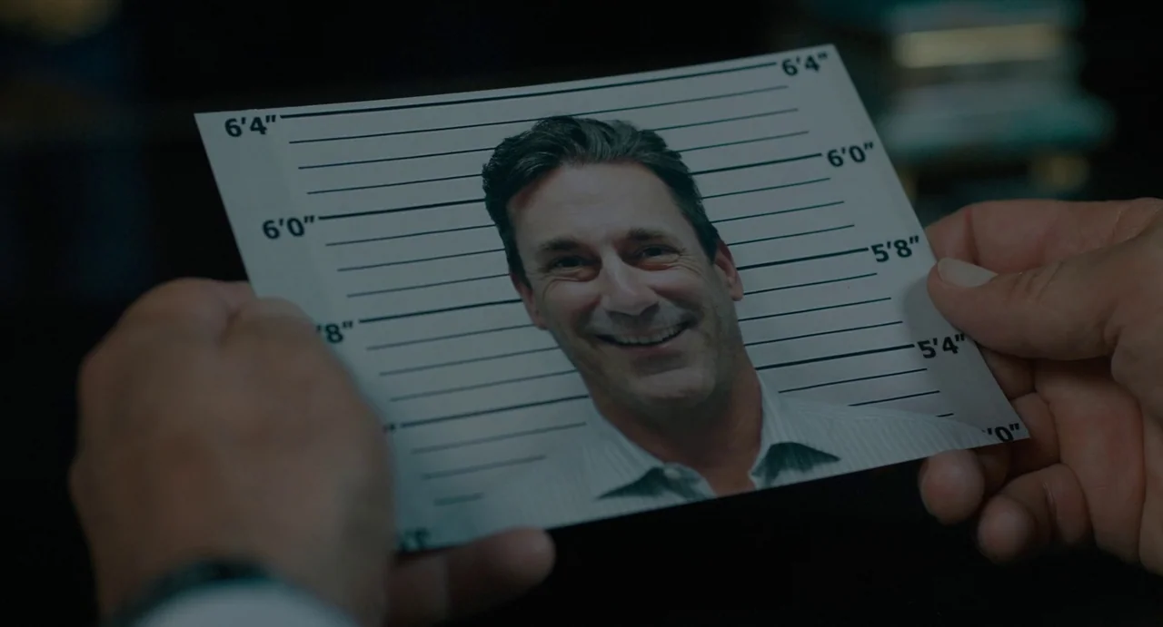 Confess, Fletch (2022) movie still: closeup — A pair of hands holds a printed mugshot-style photograph with height markers. The photo depicts a man…