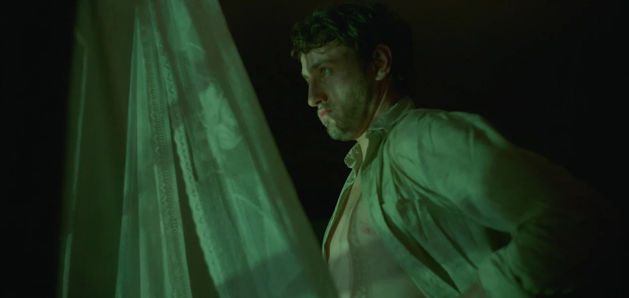 Foe (2023) movie still: medium — A dimly lit room at night features a man standing beside a draped white curtain. The space is largely obscured by…