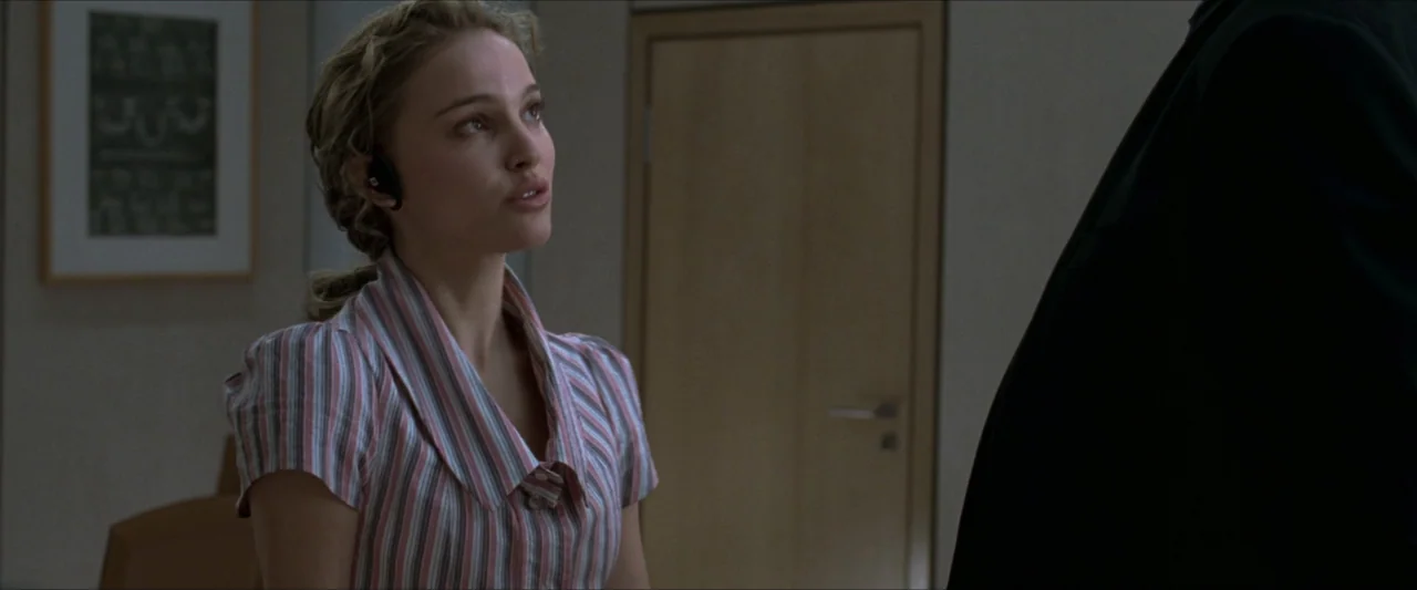 V for Vendetta (2006) movie still: medium — A sterile office hallway contains a light-colored wooden door and a framed wall print. A woman wearing a…