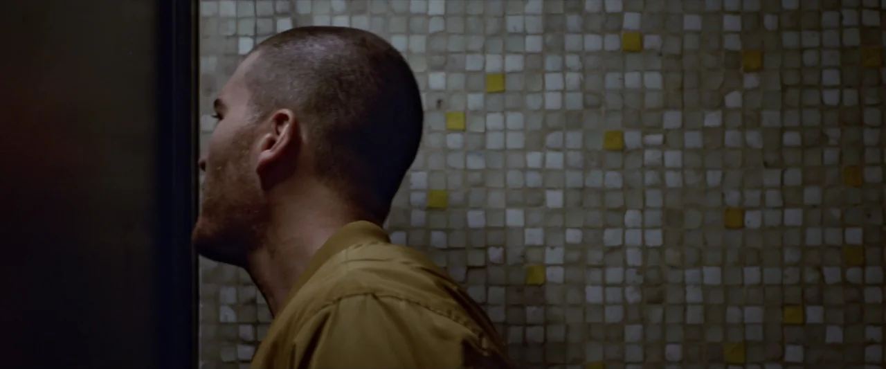 Like or Die (2023) movie still: medium — A small indoor space features a wall covered in textured square tiles. A man in a mustard yellow shirt…