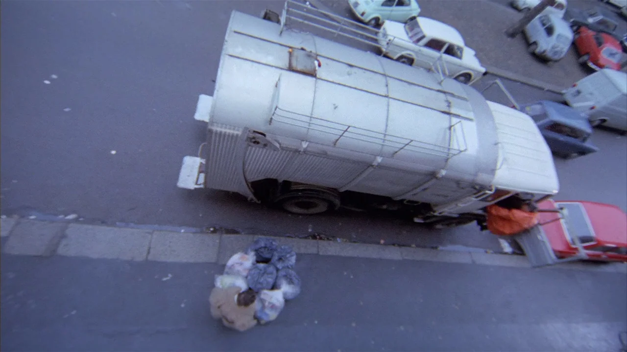 Death Occurred Last Night (1970) movie still: wide — A silver sanitation vehicle travels along an urban street with several small parked cars. Bags…
