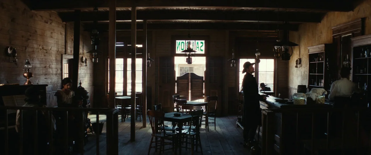 The Dead Don’t Hurt (2023) movie still: wide — A dimly lit 19th-century wooden saloon features rustic furniture and hanging oil lamps throughout. A…