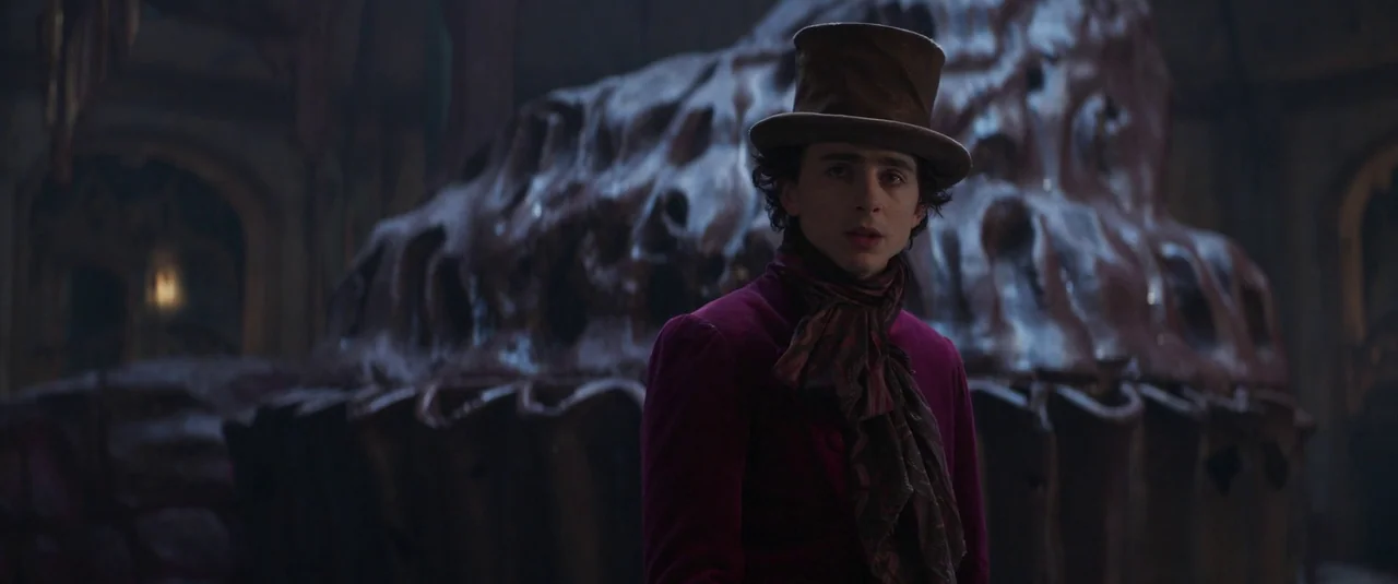 Wonka (2023) movie still: medium — A dark, dimly lit industrial space features a massive, sculptural chocolate formation behind the subject. A young…