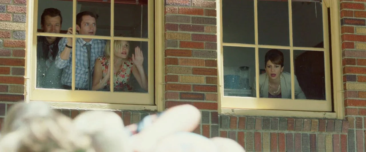 Cooties (2014) movie still: wide — A low-angle view shows the exterior of a red brick building with two windows. Four people look out from behind…