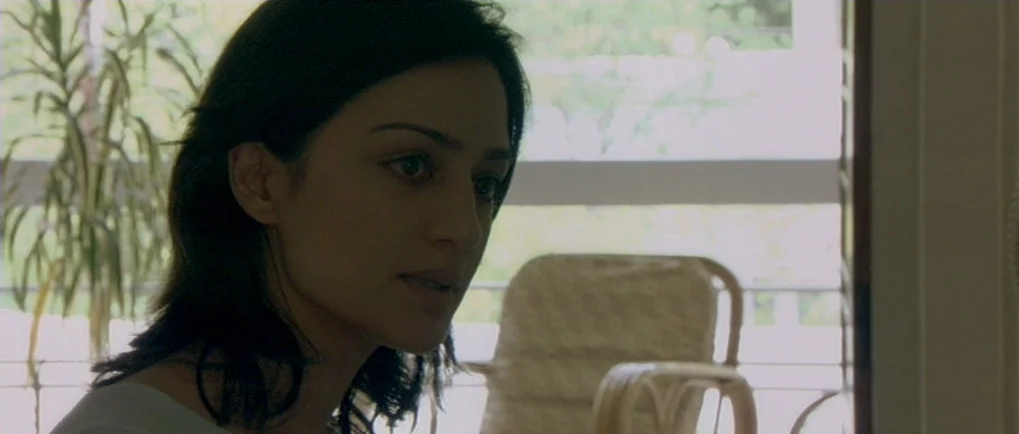 A Mighty Heart (2007) movie still: closeup — A woman stands indoors in front of a window with a sheer horizontal blind. A wicker chair is visible in…