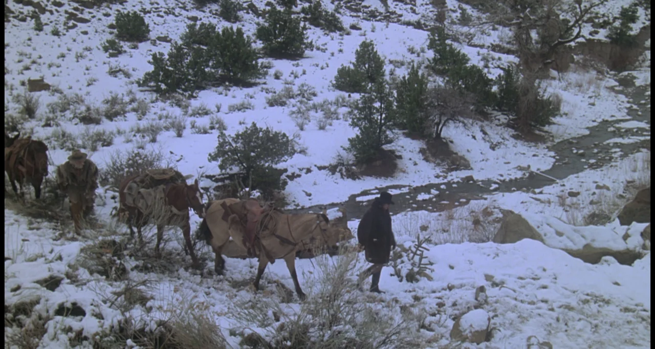 The White Buffalo (1977) movie still: wide — A snow-dusted, rocky slope features sparse shrubs and a small, shallow flowing creek. Two men lead…