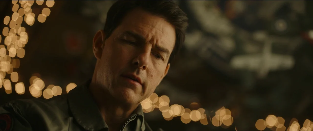 Top Gun: Maverick (2022) movie still: closeup — A dimly lit space features blurry warm lights in the background of a bar. A man in a flight suit…