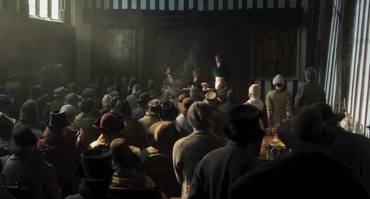 Peterloo (2018) movie still: wide — A dark, wood-paneled room is filled with a crowd of people wearing historical clothing. Three men sit at a desk…