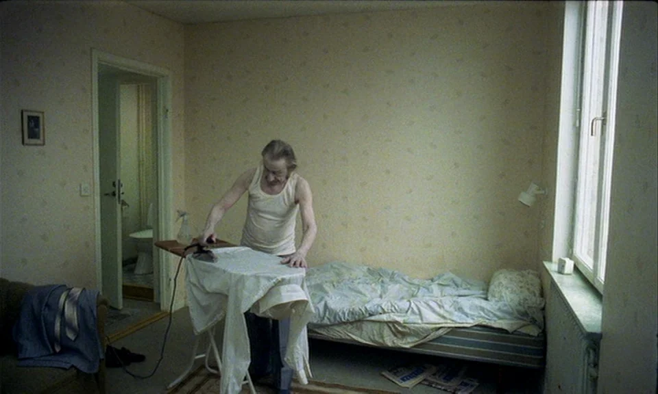 You, The Living (2007) movie still: medium — A sparsely furnished room features yellow floral wallpaper and a single bed. An elderly man in a white…