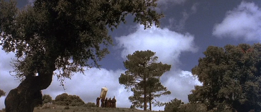 Flesh+Blood (1985) movie still: wide — A grassy hillside is framed by large gnarled oak trees against a cloudy sky. Small figures in long robes…