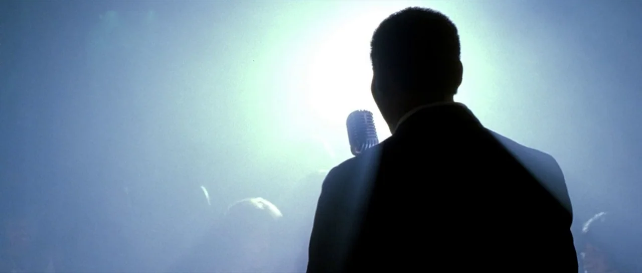 Ali (2001) movie still: medium — A dimly lit stage filled with haze and a single intense spotlight. A man in a dark suit stands with his back to the…