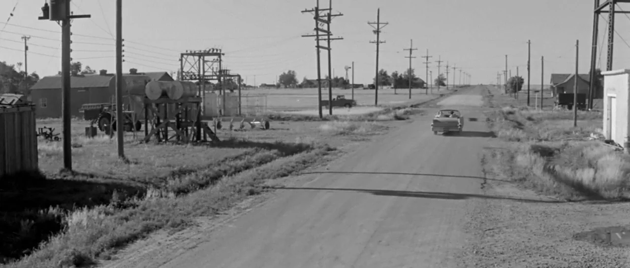 Hud (1963) movie still: wide — A desolate rural landscape features an unpaved road lined with numerous utility poles. A classic convertible car…