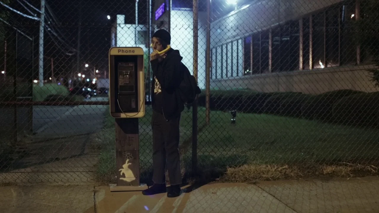 Buzzard (2014) movie still: fullbody — A night scene outside a chain-link fence separating the sidewalk from a lit building. A young person stands…