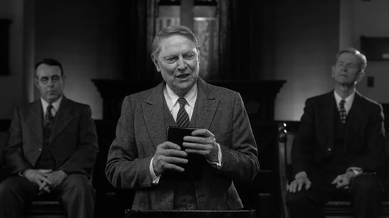 The Notorious Bettie Page (2005) movie still: medium — A small wooden pulpit stands in front of a dim room with two seated men. The central figure…