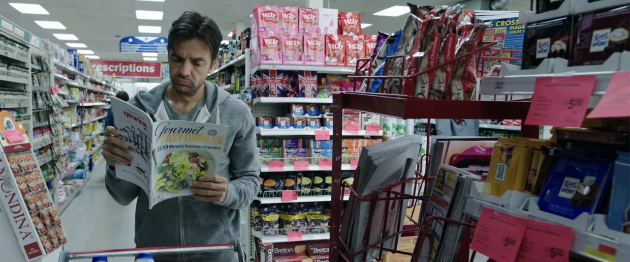 Overboard (2018) movie still: medium — A man stands in an aisle of a grocery store filled with snack items. He holds an open magazine and looks down…