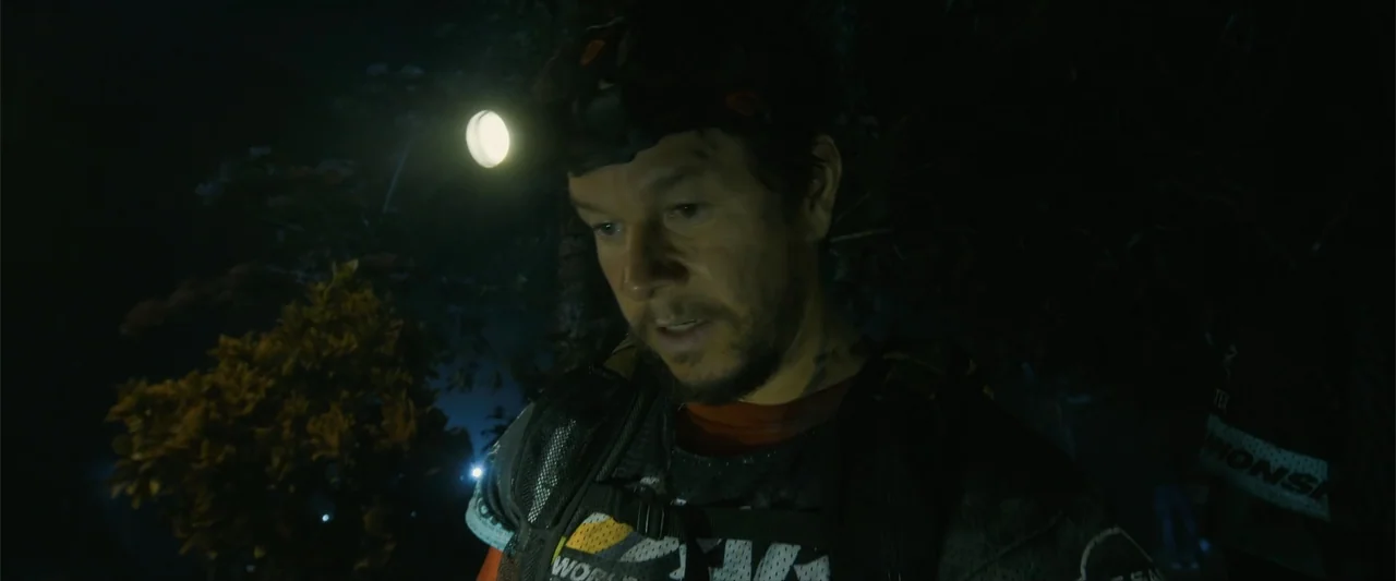 Arthur the King (2024) movie still: medium — A man stands in a dark wooded area at night. He wears a headlamp while looking down at the ground.