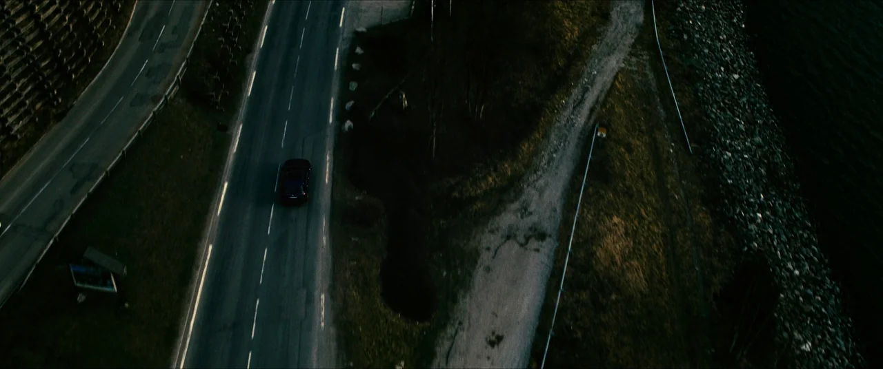 The Five Devils (2022) movie still: establishing — An aerial view of a narrow paved road bordering a dark body of water. A single dark car travels…